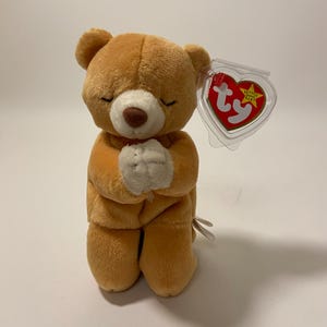 May include: A brown and white praying bear plush toy with a Ty Beanie Baby tag attached. The tag is red with a white star and the words "Beanie Babies" printed on it.