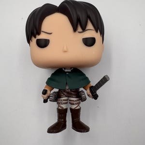 May include: A vinyl collectible figure featuring a character with black hair, a dark green cape, brown pants, and brown boots. The figure holds a grey blade in one hand and has a harness strapped around the legs and waist, with small grey utility boxes attached to the sides.