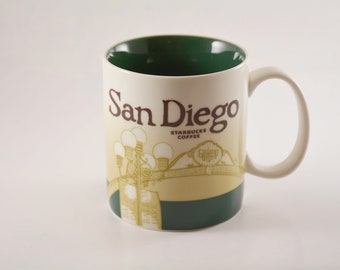 May include: A white ceramic mug with a green interior and a brown and yellow design featuring the text "San Diego" and "Starbucks Coffee". The design also includes a depiction of streetlights and a building.