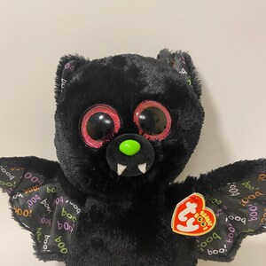 May include: Black plush bat with large pink eyes and a green nose. The bat has pink feet and wings with a "boo!" pattern. The bat has a Ty tag attached to its wing.