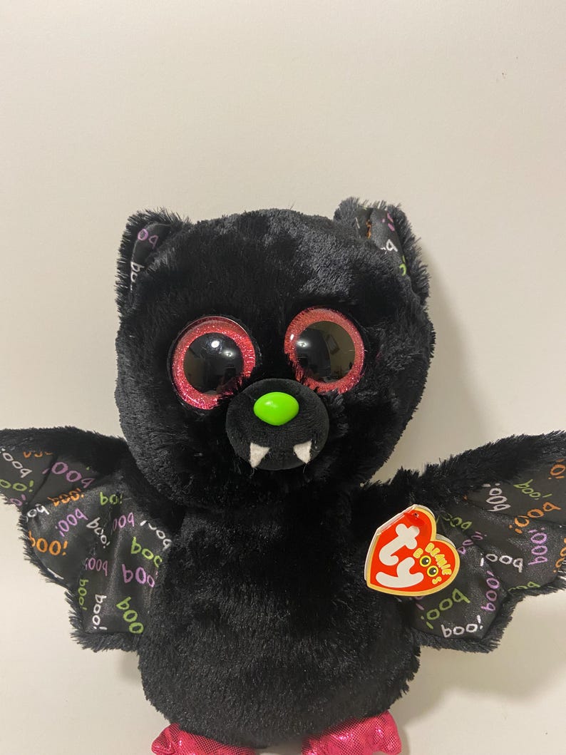 May include: Black plush bat with large pink eyes and a green nose. The bat has pink feet and wings with a "boo!" pattern. The bat has a Ty tag attached to its wing.