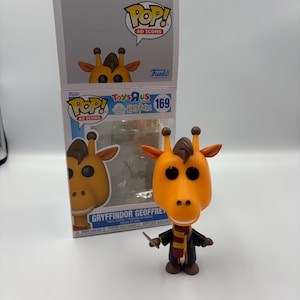 May include: A Funko Pop! vinyl figure of Geoffrey the Giraffe in a Gryffindor outfit. The figure has an orange head, brown eyes, and a black robe with a red and gold scarf. The figure is standing in front of its box.