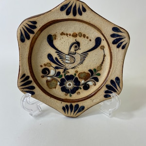 May include: A ceramic plate featuring a bird and floral design. The plate has a beige background with a brown rim and blue and brown accents. The bird is in the centre with its wings spread. The plate is supported by a clear plastic stand.