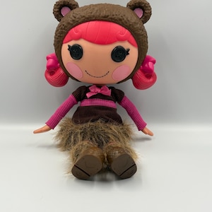 May include: A seated doll wearing a bear-themed hat and outfit. The doll has a light skin tone, pink hair, and black button eyes. The outfit includes a pink and brown striped top, a brown skirt, and furry brown boots.