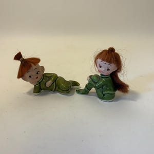 May include: Two ceramic elf figurines. One is lying down, the other is sitting with knees bent. Both wear green outfits and have red hair. The figurines have painted faces with rosy cheeks. They are on a white surface.