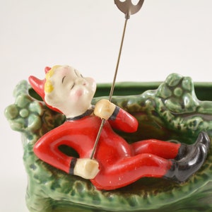 May include: A green ceramic planter shaped like a leafy branch with a red devil figurine sitting on top. The devil is holding a metal rod with a decorative top.