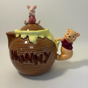 May include: A brown ceramic teapot with the word "HUNNY" in raised letters. The lid is topped with a pink Piglet figurine, and Winnie the Pooh is on the handle. The teapot has a yellow honey-like design.