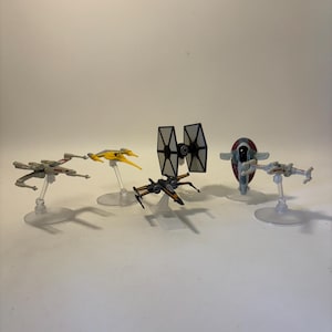May include: A collection of Star Wars miniature spacecraft toys, including X-wings, a TIE fighter, and Slave I, displayed on clear stands. The models feature detailed paint schemes with various colors, set against a plain background.