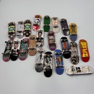 May include: A collection of twenty-two miniature finger skateboards arranged in a group. Each fingerboard features unique graphic designs, including portraits, floral patterns, skulls, and brand logos, with metal trucks and small plastic wheels.