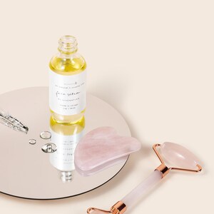 May include: A set of rose quartz beauty tools for facial massage. The set includes a roller and a gua sha tool. The roller has a rose quartz stone on a white handle with gold accents. The gua sha tool is a heart-shaped rose quartz stone. A bottle of face serum is also included. The bottle is clear glass with a white label that says "face serum rejuvenating made in korea 30 ml 1 oz."