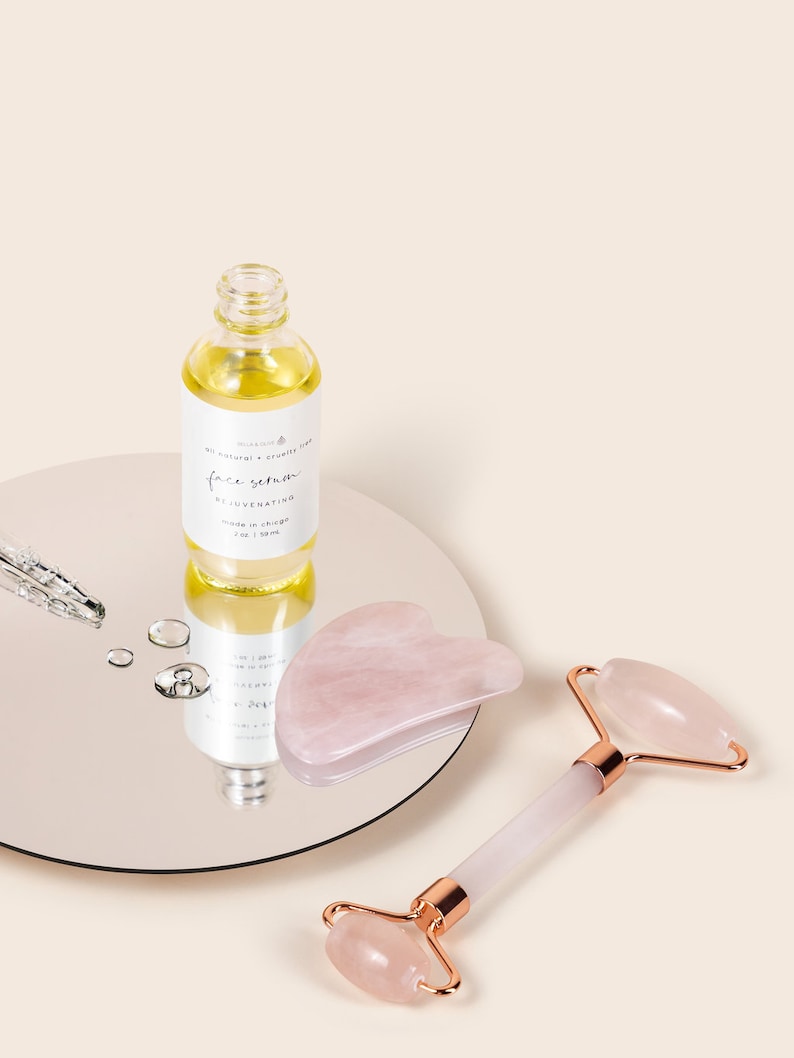 May include: A set of rose quartz beauty tools for facial massage. The set includes a roller and a gua sha tool. The roller has a rose quartz stone on a white handle with gold accents. The gua sha tool is a heart-shaped rose quartz stone. A bottle of face serum is also included. The bottle is clear glass with a white label that says "face serum rejuvenating made in korea 30 ml 1 oz."