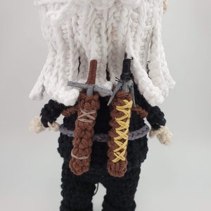 May include: Crocheted doll of a character with white hair and a white cloak. The doll wears a black outfit with two brown sword sheaths on its back. The swords are gray and the sheaths are tied with brown and yellow yarn.