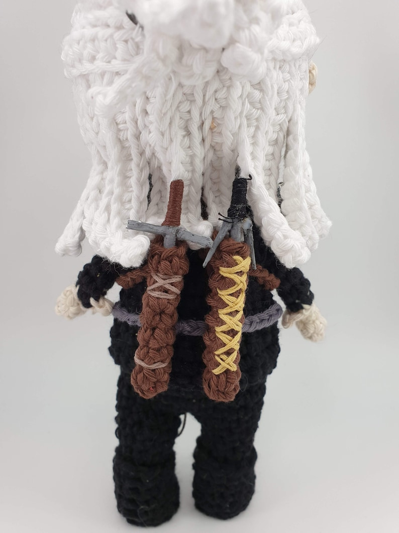 May include: Crocheted doll of a character with white hair and a white cloak. The doll wears a black outfit with two brown sword sheaths on its back. The swords are gray and the sheaths are tied with brown and yellow yarn.