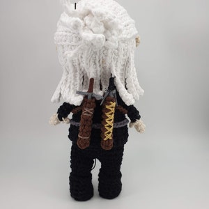 May include: Crocheted doll of a character with white hair and black clothing. The doll has two swords on its back, one brown and one black with yellow accents. The doll's hair is white and the doll is wearing a black outfit.