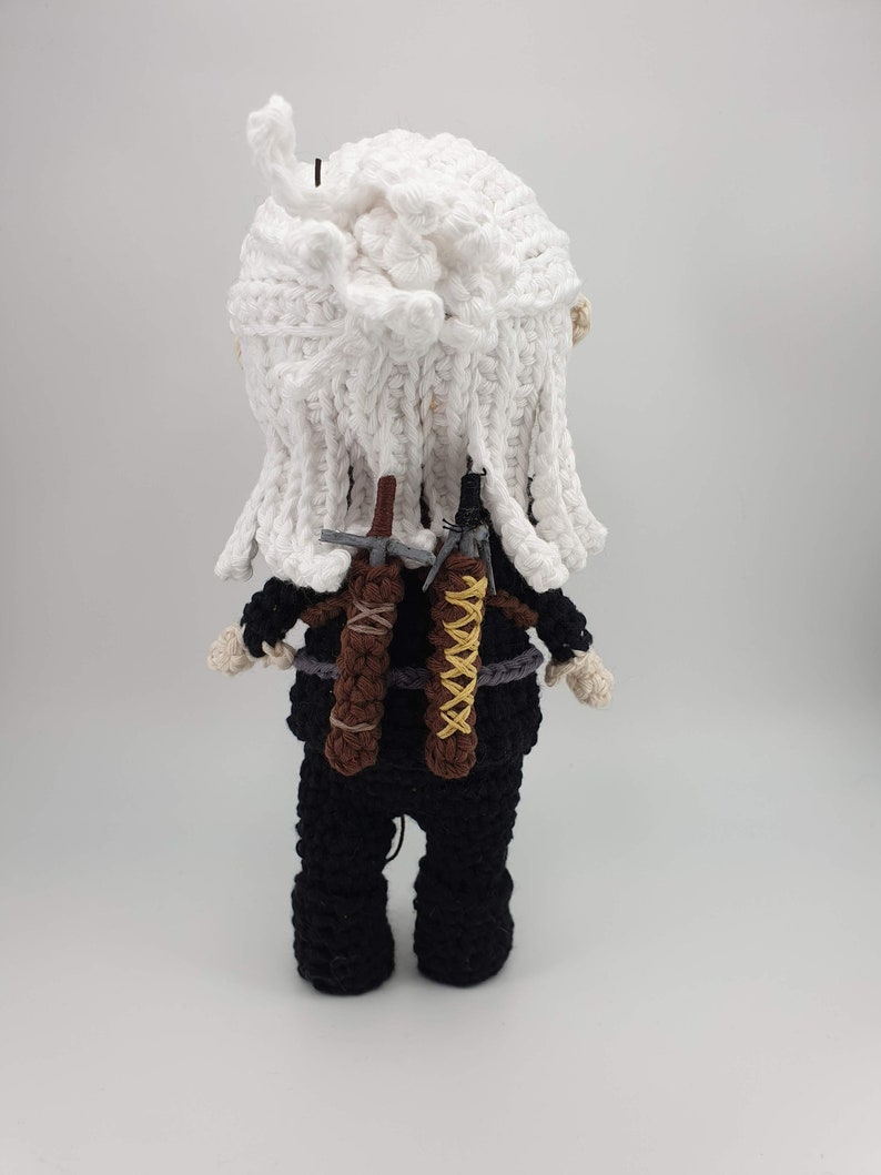 May include: Crocheted doll of a character with white hair and black clothing. The doll has two swords on its back, one brown and one black with yellow accents. The doll's hair is white and the doll is wearing a black outfit.