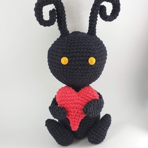 May include: A black crocheted plush toy with yellow eyes and curled antennae holding a red crocheted heart.
