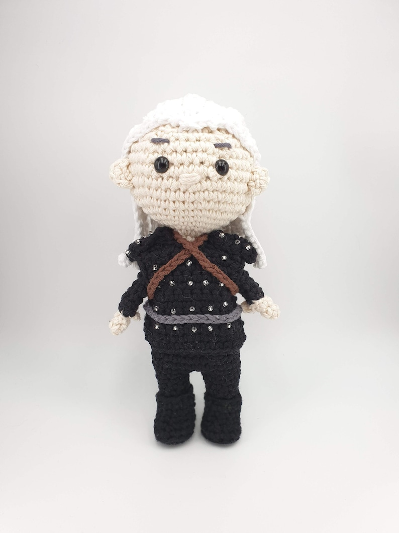 May include: A handmade crochet doll with a cream-colored head and white hair. The doll wears a black outfit with silver embellishments and a brown strap across the chest. The doll has black eyes and a neutral expression.