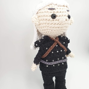 May include: Crocheted doll featuring a character with white hair and a black outfit adorned with silver embellishments. The doll has a light skin tone, black eyes, and a brown strap across the chest. The doll is standing on a white surface.
