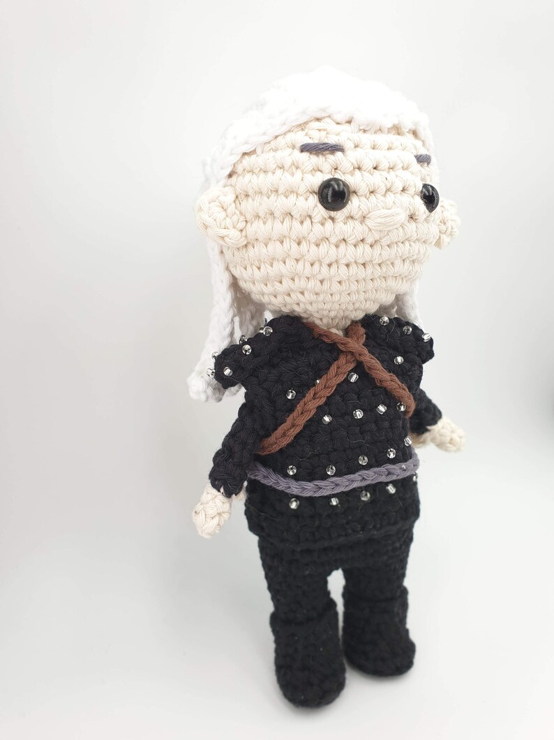May include: Crocheted doll featuring a character with white hair and a black outfit adorned with silver embellishments. The doll has a light skin tone, black eyes, and a brown strap across the chest. The doll is standing on a white surface.