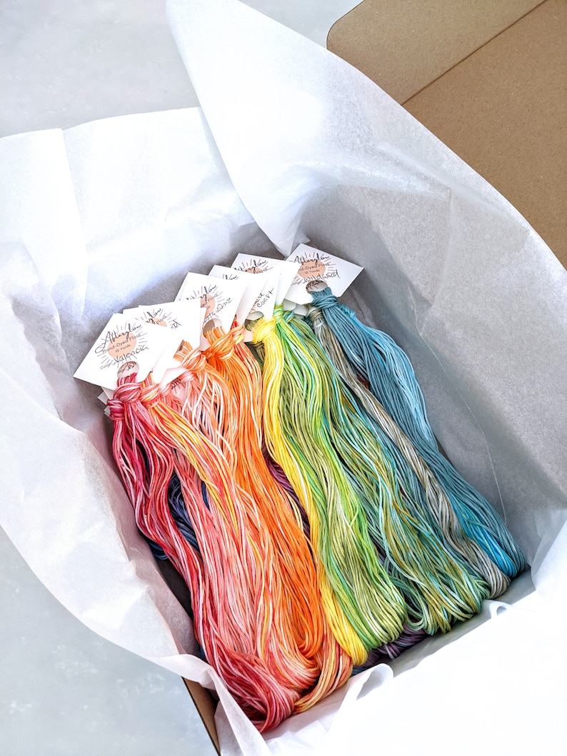 Puede incluir: Una caja llena de carretes de hilo de bordar de colores. El hilo est&aacute; dispuesto en forma de arco&iacute;ris, con cada carrete etiquetado con el nombre del color.