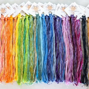 Puede incluir: Una colecci&oacute;n de 24 ovillos de hilo de bordar te&ntilde;ido a mano en un arco&iacute;ris de colores. El hilo est&aacute; dispuesto en filas, con cada ovillo etiquetado con el nombre "Allyglow Hand Dyed Floss".