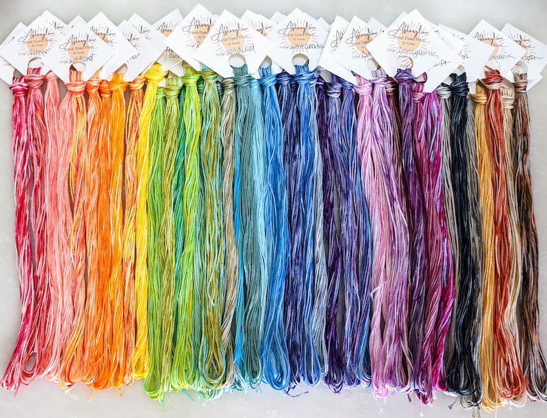 Puede incluir: Una colecci&oacute;n de 24 ovillos de hilo de bordar te&ntilde;ido a mano en un arco&iacute;ris de colores. El hilo est&aacute; dispuesto en filas, con cada ovillo etiquetado con el nombre "Allyglow Hand Dyed Floss".