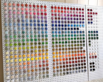 May include: A white pegboard with rows of small plastic containers filled with colorful embroidery floss. The floss is organized by color, with shades of pink, purple, blue, green, yellow, orange, red, brown, and gray.