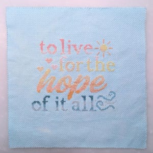 May include: A light blue cross-stitch fabric square with the words "to live for the hope of it all" in colorful thread. The design includes a sun, hearts, and decorative swirls.