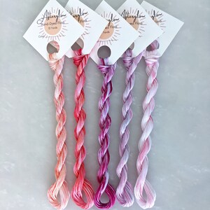 May include: Five spools of hand-dyed embroidery floss in shades of pink, orange, and purple. Each spool is labeled "Afterglow Hand-Dyed Floss 10 Yards".
