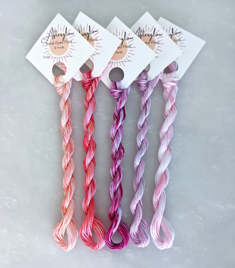 May include: Five spools of hand-dyed embroidery floss in shades of pink, orange, and purple. Each spool is labeled "Afterglow Hand-Dyed Floss 10 Yards".