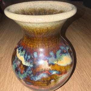 Może przedstawiać: Ceramiczny wazon z szerokim, rozszerzającym się brzegiem i zaokrąglonym korpusem. Powierzchnia pokryta jest reaktywnym szkliwem w odcieniach głębokiego brązu, bursztynu i ochry, z akcentami żywych smug w kolorze turkusowym, niebieskim i kremowym.