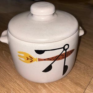 Peut inclure: Un pot en céramique crème avec couvercle, deux anses latérales et une finition mouchetée. L'avant présente une illustration graphique d'une fourchette jaune et d'un ensemble de cuillères à mesurer noires avec un manche texturé marron.