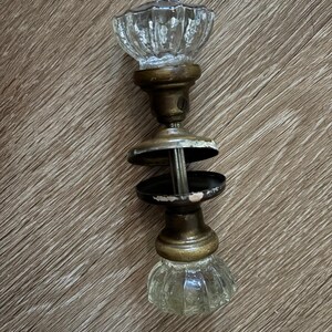 May include: A vintage door knob set featuring two clear, fluted glass knobs attached to brass-toned metal bases. The assembly includes a central metal spindle and two circular brass-toned rosettes with visible signs of wear and aged patina.