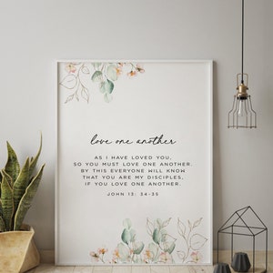 May include: A white framed print with a watercolour floral design and the text "love one another." The text continues with a bible verse from John 13:34-35: "As I have loved you, so you must love one another. By this everyone will know that you are my disciples, if you love one another."