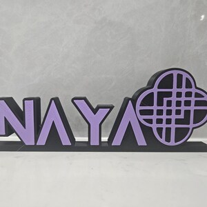May include: A black and purple decorative sign that spells out the name "NAYA". The letters are purple with black outlines, and the design includes a stylized floral shape. The sign is made of a solid material and stands upright.