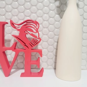May include: Pink wooden LOVE sign with a knight's helmet design in the center of the 'O'. The sign is on a white tiled background.