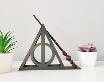 May include: A dark gray triangle with a circle inside, resembling the Deathly Hallows symbol from the Harry Potter series. A brown wand is leaning against the triangle. Two small white pots with green and purple plants are on either side of the triangle.