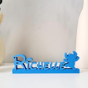 May include: Blue wooden name sign with the name "Richelle" and a blue Stitch character from Lilo & Stitch.