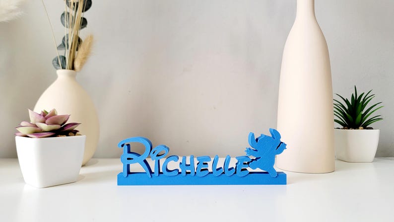 May include: Blue wooden name sign with the name "Richelle" and a blue Stitch character from Lilo & Stitch.