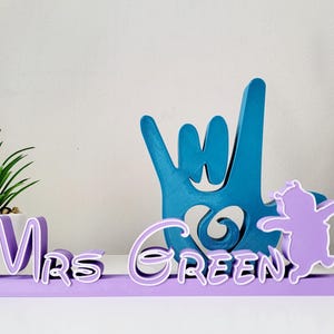 May include: Purple wooden sign with the text "Mrs. Green" in a cursive font. The sign also features a blue hand making the "I love you" sign language gesture and a purple silhouette of a cartoon character.