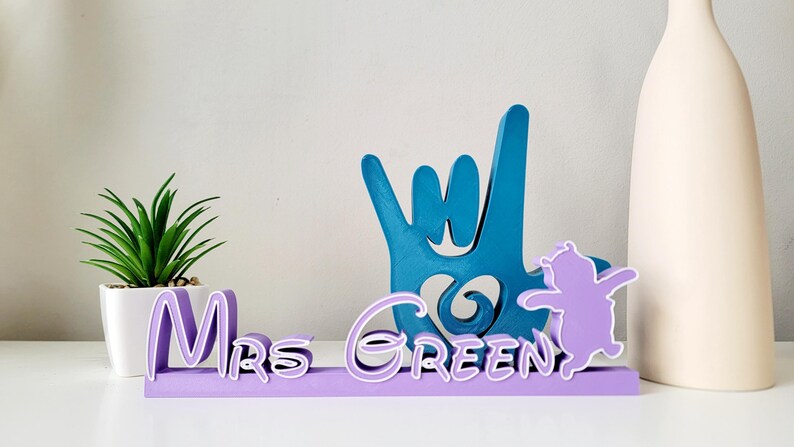 May include: Purple wooden sign with the text "Mrs. Green" in a cursive font. The sign also features a blue hand making the "I love you" sign language gesture and a purple silhouette of a cartoon character.