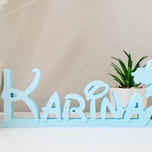 May include: Light blue wooden name sign with the name "Karina" spelled out in a whimsical font. A mermaid silhouette is on the right side of the sign.