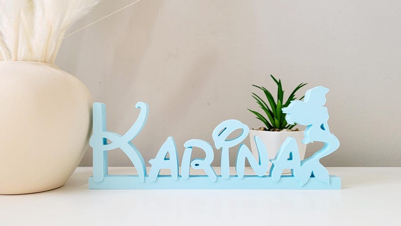 May include: Light blue wooden name sign with the name "Karina" spelled out in a whimsical font. A mermaid silhouette is on the right side of the sign.