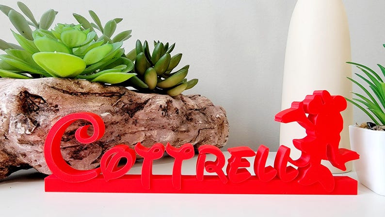 May include: Red 3D printed name sign with the name "COTRELL" in a stylised font. The sign is sitting on a wooden surface with green succulents in the background.