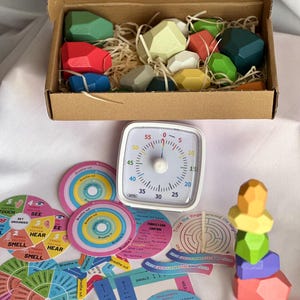 Puede incluir: Un conjunto de herramientas de mindfulness, que incluye bloques de madera de colores en una caja de cartón, un temporizador y varias tarjetas con texto y gráficos. Los bloques son de color verde, rojo, azul y amarillo. El temporizador tiene una esfera blanca con números negros.