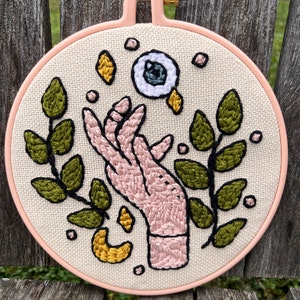 May include: Embroidered hoop art featuring a pink hand reaching towards a white eye with a blue iris. The hand is surrounded by green leaves and yellow crescent moons.
