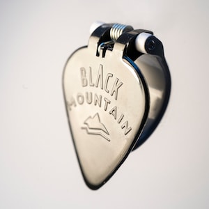 May include: A silver guitar pick holder with a spring mechanism. The pick holder is shaped like a guitar pick and has the words "BLACK MOUNTAIN" engraved on it.