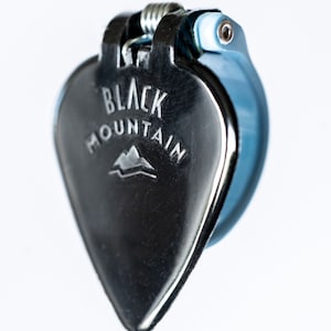 May include: A silver guitar pick with a blue clamp. The pick has the words "Black Mountain" and a mountain logo engraved on it.