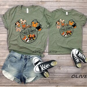May include: Two olive green t-shirts with a Mickey Mouse head design featuring autumn-themed elements like pumpkins, coffee cups, and a car. The text "Fall Vibes" is inside the Mickey Mouse head. The shirts are on a wooden surface with a pair of black Converse shoes.