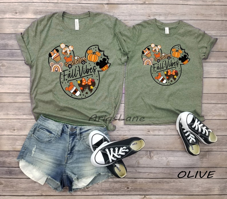 May include: Two olive green t-shirts with a Mickey Mouse head design featuring autumn-themed elements like pumpkins, coffee cups, and a car. The text "Fall Vibes" is inside the Mickey Mouse head. The shirts are on a wooden surface with a pair of black Converse shoes.